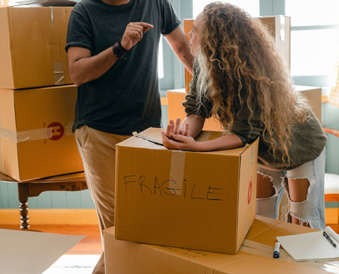 Girl leaning on a cardboard box that has "Fragile" written on it. She is talking to someone stood next to her.