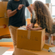 Girl leaning on a cardboard box that has "Fragile" written on it. She is talking to someone stood next to her.